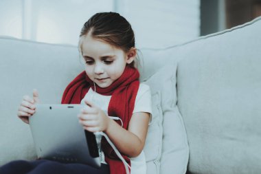 Tablet kullanarak soğuk ile kırmızı fularlı küçük kız. Genç kız hasta. Oda beyaz kanepe. Mutsuz çocuk. Hastalık kavramı. Sağlık ve sağlıklı yaşam konsepti. Tablet Pc ile küçük kız.