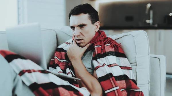 Man with Cold Sitting on Sofa in Checkered Blanket. Man at Home. Sick Young Man. White Sofa in Room. Unhappy Guy. Disease Concept. Healthcare and Healthy Lifestyle Concept. Using Laptop.