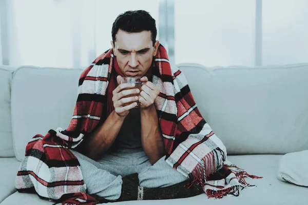 Man with Cold Sitting on Sofa in Checkered Blanket. Man at Home. Sick Young Man. White Sofa in Room. Unhappy Guy. Disease Concept. Healthcare and Healthy Lifestyle Concept. Drinking Medicines.