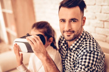 Oturun. Joystick elinde. Sanal gerçeklik maskesi. Baba ve kızı. Birlikte vakit geçirmek. Babalar günü. Adam oyun kız ile. Video oyunu. Beyaz iç. Koltukta oturuyor. Kız sanal maskede görünüyor