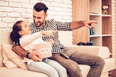 Baba ve kızı. Birlikte vakit geçirmek. Babalar günü. Kızla oynayan adam. Video oyunu. Beyaz iç. Elinde patlamış mısır. Patlamış mısır yemek. Patlamış mısır ile kanepe üzerinde durur. Parmak ile işaret. Ekose gömlek.