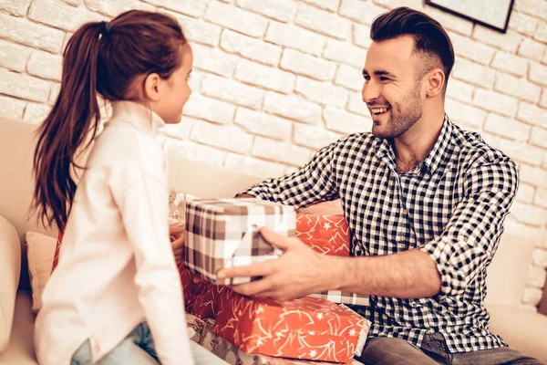 Man Gives Lot Gift Boxes. Spend Time Together. Father\'s day. Man Play with Girl. Smiling Girl at Home. Sit on Sofa. Gift box. Gift in Red Pack. Man with Gift for Child. Make Surprise for Daughter.