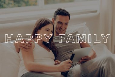 Yatakta yatarken çocuklar. Gri Tablet ebeveyn gözetiminde. Veliler konuşmak. Mutlu birlikte. Göbek kız beklenti gebelik. Hamile kadın mutlu bir şekilde gülümseyerek. Mutlu genç aile. Hamile Sit evde.