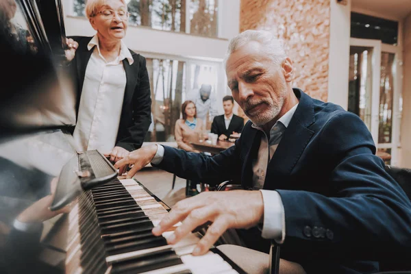 Elderly Man Plays the Piano. Doctor at Head of Table. Nursing Home. Elderly Woman Stay Near. Young People Sit at Table. Cute Relationship. Happy Holidays. Happy Together. Smiling People.