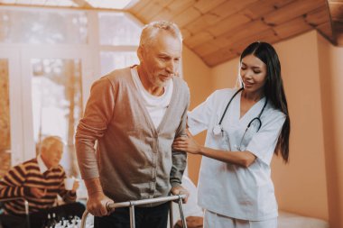 Kadın Caregiver Yaşlı kemikler. Medikal tedavi. Yetişkin beyaz sert kendisini gidin. Kadın destekler adam Asyalı hemşire yetişkin Walker yardımcı olur. Emekli go-kart üzerinde. Hemşire tutun emekli kişi. Huzurevi.