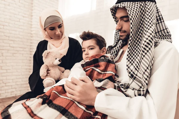 Sick Arabian Boy in Blanket on Sofa with Family. Parent with Child Indoor. Unhappy Boy. Young Arabian Woman. Muslim Family. Healthcare Concept. Checkered Blanket. Boy with Flu. White Sofa.