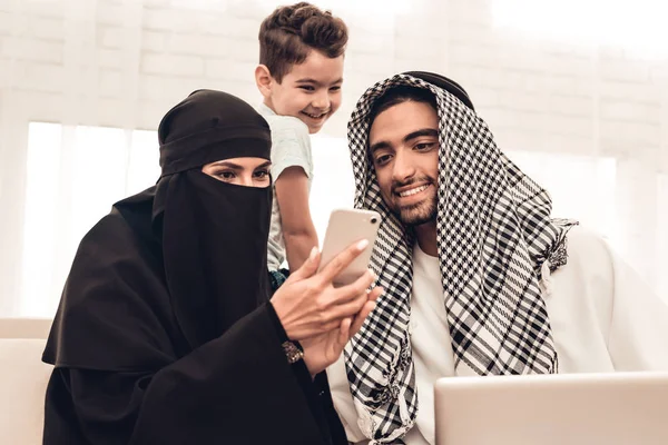 年轻的阿拉伯家庭在家里使用沙发上的笔记本电脑 家庭坐在沙发上 使用数字设备的人 微笑的男孩在家里 穆斯林家庭 微笑的男孩 年轻的阿拉伯女人 黑面纱中的妇女 — 图库照片