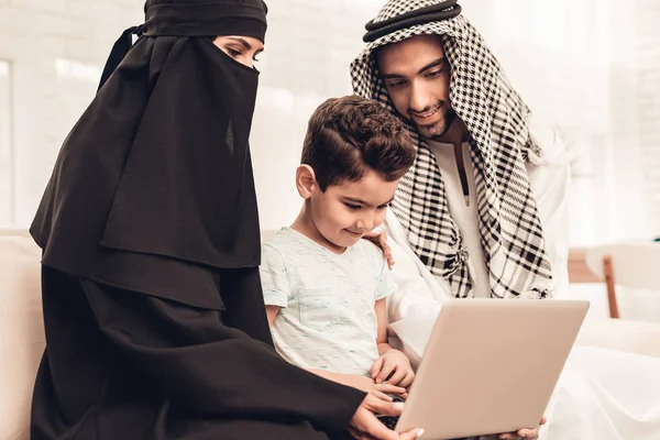 年轻的阿拉伯家庭在家里使用沙发上的笔记本电脑 家庭坐在沙发上 使用数字设备的人 微笑的男孩在家里 穆斯林家庭 微笑的男孩 年轻的阿拉伯女人 黑面纱中的妇女 — 图库照片