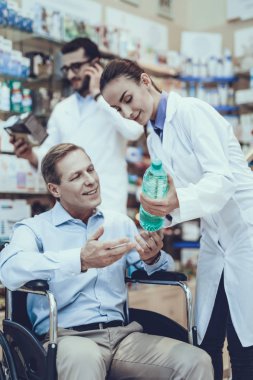 Kadın eczacı yardımcı olur istemciye bir maden suyu seçin. Tekerlekli sandalyede oturan istemci. Erkek eczacı smartphone üzerinde konuşuyor. Eczacılar Weares özel tıbbi üniforma. Eczanede bulunan insanlar.