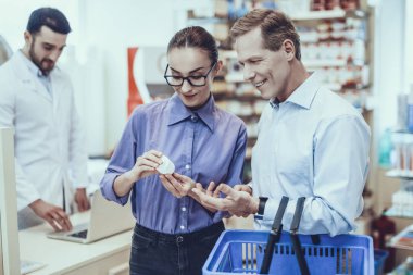 Müşteriler bir parfümden seçin. Müşteriler yetişkin erkek ve genç kadın var. Bir şişe hap ile tutan kadın. Bir alışveriş sepeti tutan adam. Eczacı arka plan üzerinde bir Laptop kullanıyor. İnsanlar eczane.