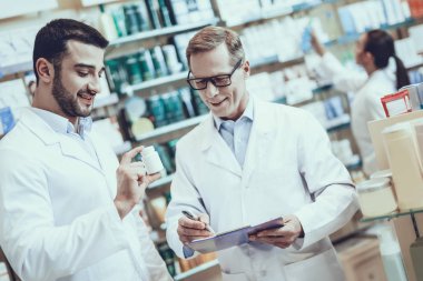 İki Eczacılar çalışıyor. Eczacılar beyaz yetişkin adam ve genç adam. Bir şişe hap ile gösterilen adam. Adam bir ilaç hakkında bilgi yazma. Eczanede bulunan insanlar.