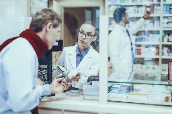 Illness Man Buys a Medicament. Female Pharmacist Propose a Bottle with Pills to Man. Male Pharmacist Choose a Medicament on Background. Pharmacists is Wearing a Medical Uniform. People in Pharmacy.