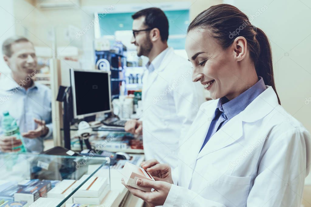 Pharmacists Working at Counter. Man Buys a Mineral Water at Male Pharmacist. Female Pharmacist Holding a Box with Medicaments. Pharmacists Wearing a Medical Uniform. People Located in Pharmacy.