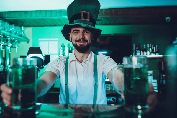 Saint Patrick\'s Day Party. Barman is Holding a Traditional Green Beer. Barman is Young Smiling Beard Man. Man Standing at Bar Counter. Man Wearing a Green Hat. Pub Interior. Green Tone.