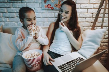 Anne ile kızı koltukta oturuyor. İnsanları Laptop hakkında bir Video izlemek. Patlamış mısır yemek kişi. Anne ve kızı gülen var. Duvar resim arka plan ile. Akşam saat. Ev iç.