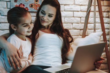 Anne ile kızı koltukta oturuyor. İnsanları bir dizüstü bilgisayarda bir şey hakkında bir Video izlemek. Anne ve kızı gülen var. Duvar resim arka plan ile. Akşam saat. Ev iç.