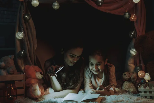 母女躺在假帐篷下的地毯上 母亲和女儿正在看书 母亲用手机在书上发光 人是微笑 晚上的时间 家居内饰 — 图库照片