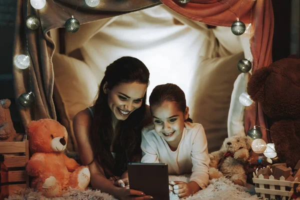 母女躺在地毯上 躺在假帐篷下的人 母女们正在平板电脑上观看视频 人们快乐而微笑 晚上的时间 家庭内部的人 — 图库照片