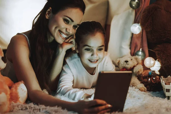 母女躺在地毯上 躺在假帐篷下的人 母女们正在平板电脑上观看视频 人们快乐而微笑 晚上的时间 家庭内部的人 — 图库照片
