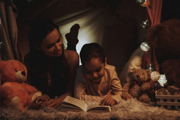 母女躺在地毯上 躺在假帐篷下的人 女儿正在给妈妈读一本书 人是快乐和微笑的 晚上的时间 家庭内部的人 — 图库照片