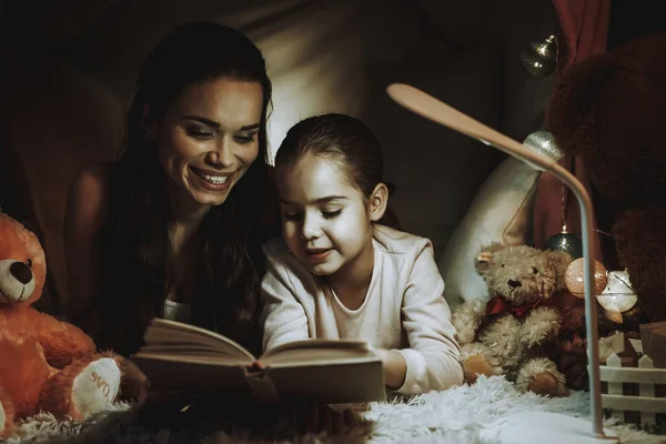 母女躺在地毯上 躺在假帐篷下的人 母亲和女儿正在看灯下的一本书 人是快乐和微笑的 晚上的时间 家庭内部的人 — 图库照片