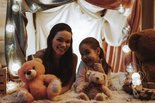母女躺在假帐篷下的地毯上 母亲和女儿抱着泰迪熊 人是快乐和微笑的 女孩显示好的标志 晚上的时间 家居内饰 — 图库照片