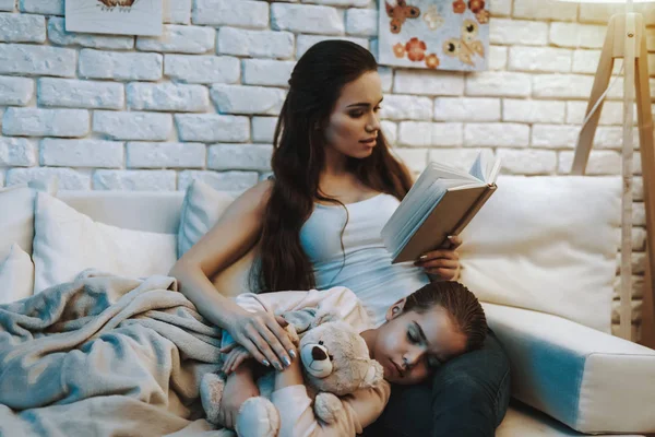 母亲在沙发上和看书 女儿睡在母亲的膝盖上 女儿抱着泰迪熊 母亲抚摸女儿和微笑 晚上的时间 家庭内部的人 — 图库照片