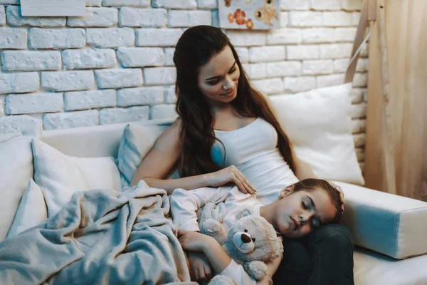 年轻的母亲坐在沙发上 女儿睡在母亲的膝盖上 女儿抱着泰迪熊 母亲抚摸女儿和微笑 晚上的时间 家庭内部的人 — 图库照片