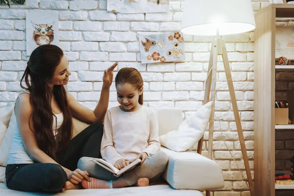 Mother with Daughter is Sitting on Couch. Daughter is Reading a Book. Mother Stroking Her Daughter. Persons is Smiling. Wall with Pictures on Background. Evening Time. Home Interior.