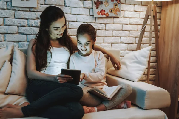 带着女儿的母亲坐在沙发上 妈妈在平板电脑上给女儿看东西 女儿拿着一本书 人是微笑 带图片的墙 晚上的时间 家居内饰 — 图库照片