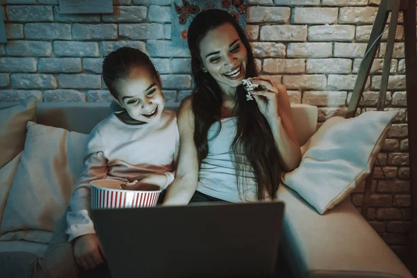 Mother with Daughter is Sitting on Couch. People Watching a Video on Laptop. Persons Eating a Popcorn. Mother and Daughter is Smiling. Wall with Pictures on Backgound. Evening Time. Home Interior.