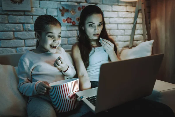 Mother with Daughter is Sitting on Couch. People Watching a Video on Laptop Computer. Persons Eating a Popcorn. Mother is Surprised. Girl is Smiling. Wall with Pictures. Evening Time. Home Interior.