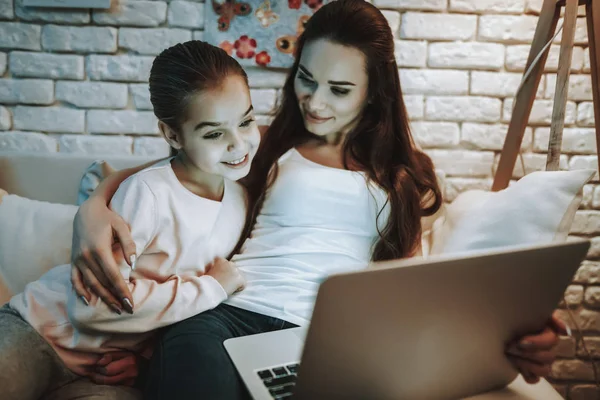 Mother with Daughter is Sitting on Couch. People Watching a Video about a Something on Laptop Computer. Mother and Daughter is Smiling. Wall with Picture on Background. Evening Time. Home Interior.