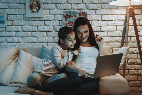带着女儿的母亲坐在沙发上 在笔记本电脑上观看视频的人 女孩指向屏幕上的母亲 人们都在微笑 背景上有图片的墙 晚上的时间 家居内饰 — 图库照片