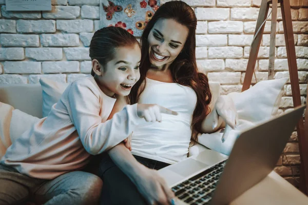 带着女儿的母亲坐在沙发上 在笔记本电脑上观看视频的人 女孩指向屏幕上的母亲 人们都在微笑 背景上有图片的墙 晚上的时间 家居内饰 — 图库照片