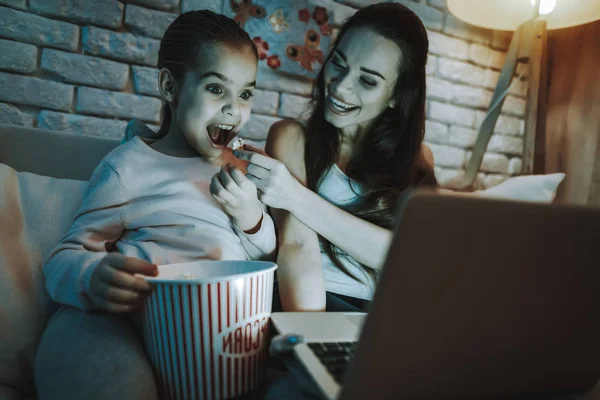 Mother with Daughter is Sitting on Couch. People Watching a Video on Laptop. Mother Feeding a Daughter a Popcorn. Mother and Daughter is Smiling. Wall with Pictures. Evening Time. Home Interior.