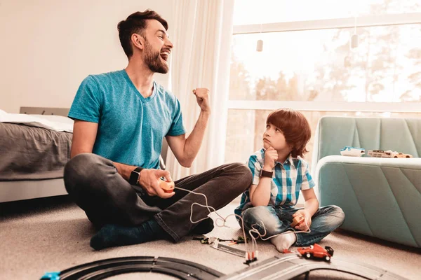 Bearded Father and Son Playing with Toy Race Road. Man Sitting on Floor. White Carpet in Room. Toy Cars. Exited Boy. Happy Family Concept. White Carpet. Lying on Floor. Indoor Fun.