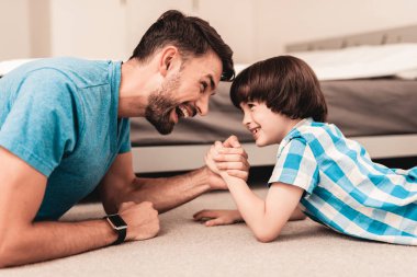 Father and Son Lying on Floor and Have Fun at Home. Sitting Boy. Young Father. Young Bearded Man at Home. Gray Sofa in Room. Happy Family Concept. White Carpet. Lying on Floor. Indoor Fun. clipart