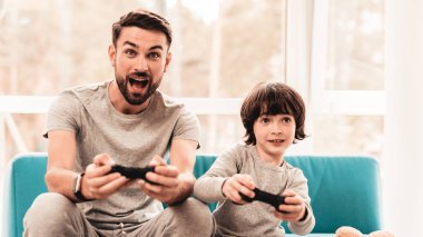 Baba ve oğul oturan ve konsolda oyun. Oturan çocuk. Genç baba. Konsolda oyun. Joystick elinde. Genç adam evde sakallı. Mavi koltukta oturuyor. Mutlu aile kavramı.