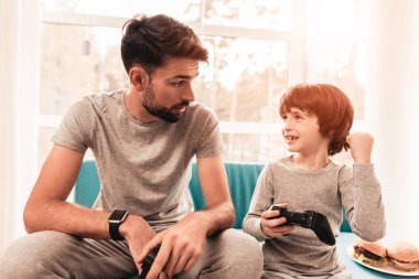 Baba ve oğul oturan ve konsolda oyun. Oturan çocuk. Genç baba. Konsolda oyun. Joystick elinde. Genç adam evde sakallı. Mavi koltukta oturuyor. Mutlu aile kavramı.