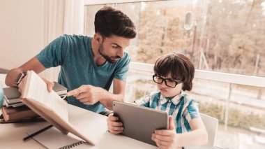 Baba oğlu gözlük ile ev ödevi yardım etmeye çalışıyorum. Evde eğitim. Oda beyaz tablosunda. Dijital Divece kullanarak çocuk. Genç baba. Evde ders. Eğitim kavramı. Gömlekli çocuk. Tablet üzerinde oynama.