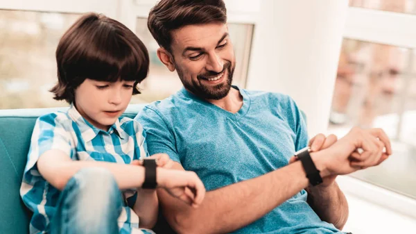 Little Boy with Father Wacthing on Hand Watches. Sitting Boy. Young Father. Young Bearded Man at Home. Sitting on Blue Sofa. Happy Family Concept. Time Concepts. Modern Hand Watch.
