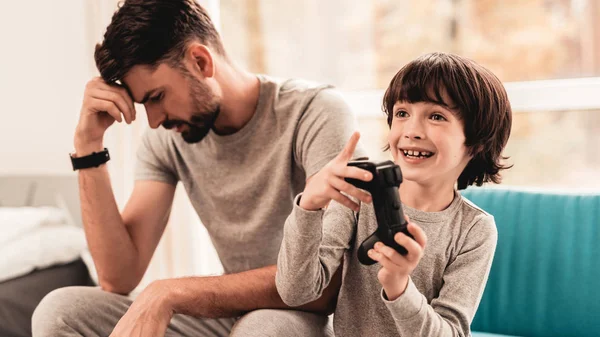 Father and Son Sitting and Playing on Console. Sitting Boy. Young Father. Playing on Console. Joystick in Hands. Young Bearded Man at Home. Sitting on Blue Sofa. Happy Family Concept.