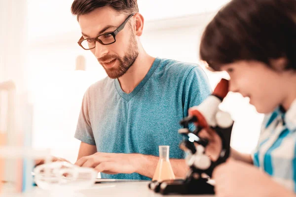 Youn Bearded Father Teaching Son in Shirt at Home. Education at Home. Using Microscope. Studding Chemistry. White Table in Room. Sitting Boy. Young Father. Lesson at Home. Education Concept.