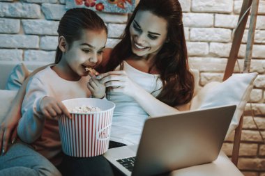 Anne ile kızı koltukta oturuyor. İnsanları Laptop hakkında bir Video izlemek. Kız bir patlamış mısır yemek. Anne ve kızı gülen var. Duvar resim arka plan ile. Akşam saat. Ev iç.