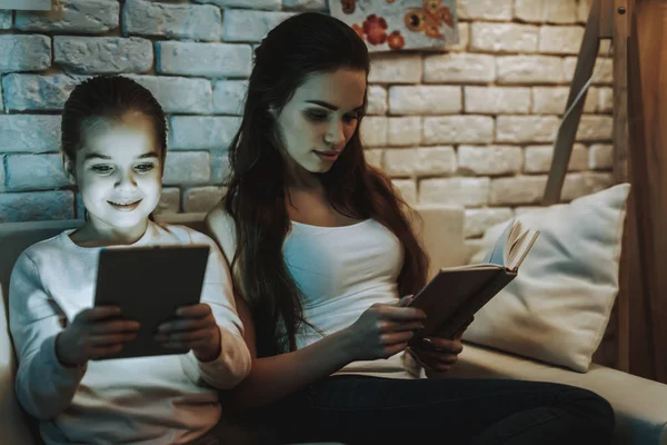 带着女儿的母亲坐在沙发上 妈妈正在看书 女儿在平板电脑上看东西 女孩在微笑 背景上有图片的墙 晚上的时间 家居内饰 — 图库照片