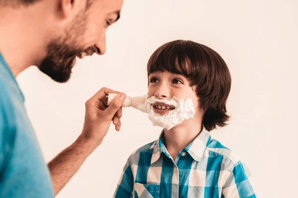 若い父は 家でひげをそるの幼い息子を教えています 顔に泡 シェービング ツールです かみそりの刃で 幸せな家族の概念 シャツの少年 衛生とスキンケア概念 衛生的な石鹸 — ストック写真