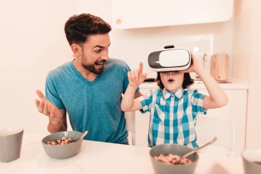 Genç baba ve oğul Vr gözlükle evde. Boy gülümseyerek. Modern mutfak. Oturan çocuk. Gömlekli çocuk. Sabah kahvaltı. Beyaz masa mutfak. Dijital aygıt kullanarak. Mutlu bir aile.