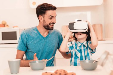 Genç baba ve oğul Vr gözlükle evde. Boy gülümseyerek. Modern mutfak. Oturan çocuk. Gömlekli çocuk. Sabah kahvaltı. Beyaz masa mutfak. Dijital aygıt kullanarak. Mutlu bir aile.