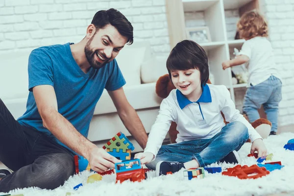 彼の息子との時間を費やしています 父親は子育てに取り組んでいます 父と長男は カーペットの上のおもちゃで遊んでいます 次男は背景に単独で再生します 寝室にあります — ストック写真